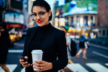 Portrait of confident businesswoman with trendy red lips sending messages on mobile while standing on street with coffee to go, prosperous female owner checking notification on smartphone outdoors