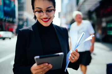 Cheerful woman in trendy eyeglasses reading positive email from friend using 4g wireless internet and application on modern digital tablet for chatting online during way to work in business company