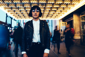 Half length portrait of charming hipster girl in stylish casual wear posing at camera in illuminated night downtown, brunette young woman in eyewear and trendy apparel in crowded time lapse city