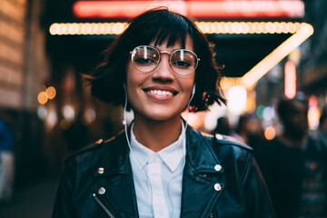 Half-length portrait of cheerful hipster girl spending time on night street of New York listening music in earphones, gorgeous young woman looking at camera enjoying favorite playlist at evening city
