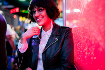 Smiling hipster girl in eyewear enjoying sound of favorite song from playlist in earphones standing...