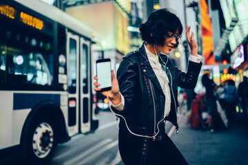 Trendy dressed young woman feeling carefree dancing to favorite song listening to in earphones, happy hipster girl having fun on evening city street with neon illumination moving to sound on music - obrazy, fototapety, plakaty