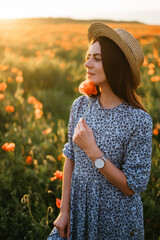  Amazing young woman in hat and blue dress posing in a poppies field. Beautiful happy tender young woman enjoying the smell of summer wildflowers