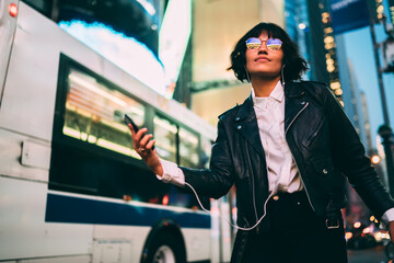 Carefree woman in fashionable clothing and spectacles with neon reflection listening tranquil music for relaxing in evening city, dancing teenager with modern smartphone technology recreating outdoors
