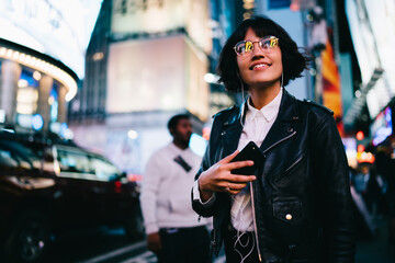 Prosperous female party girl in electronic headphones listening positive music at evening metropolitan street, happy millennial in classic eyeglasses hanging out in night downtown of New York City