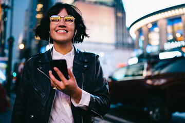 Cheerful millennial blogger with modern cellphone technology for web chatting recreating at urban street, happy generation z in electronic headphones listening positive music playlist and smiling