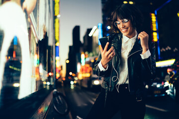 Cheerful hipster girl choosing song from mobile playlist enjoying nightlife in New York,happy trendy woman in spectacles reading message on smartphone listening music on street with neon illumination