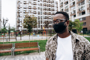 Close-up portrait of a stylish African American guy stands alone in the street in a black protective mask on his face. Precautionary measures