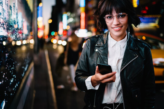 half length portrait of stylish hipster girl in spectacles and earphones smiling at camera while listening song from music app installed on phone during night walk in metropolis with neon illumination