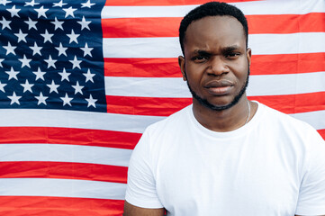Sad African American guy in a white t-shirt stands against the background of the flag of United States of America