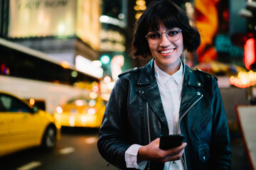 Cheerful hipster girl with mobile device smiling at metropolitan street enjoying nightlife, half length portrait of female in earphones holding smartphone using media application with music playlist