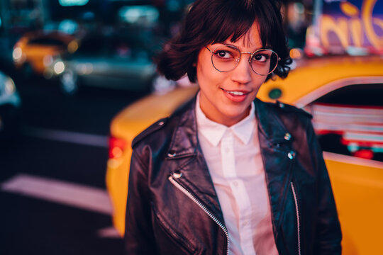 Carefree brunette young woman with short haircut in stylish eyewear for eyes protection fascinated with New York urbanity in Manhattan,youthful female in leather jacket enjoying metropolitan nightlife