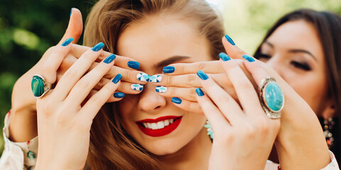 Two beautiful women playing and brunette holding hands and hiding face to her blonde girlfriend. Female with trendy stylish manicure in blue and green tones, fashion accessories, jewelry,big earrings. - obrazy, fototapety, plakaty