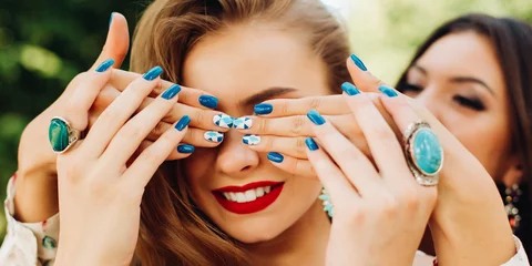  Two beautiful women playing and brunette holding hands and hiding face to her blonde girlfriend. Female with trendy stylish manicure in blue and green tones, fashion accessories, jewelry,big earrings. © Вячеслав Косько