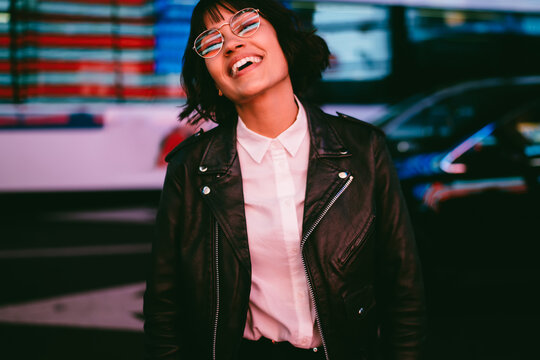 Cheerful hipster girl in spectacles with night city light reflection laughing feeling carefree on free time, smiling young woman in stylish clothing having fun in evening spending time in megalopolis.