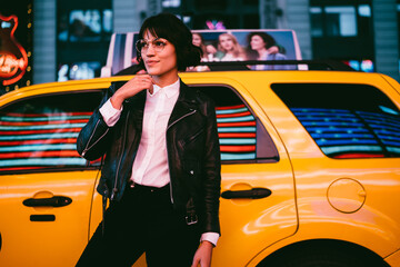 Carefree female tourist in spectacles looking away on metropolitan street thinking about evening sightseeing in New York, youthful hipster girl standing at city urbanity with cab taxi on background