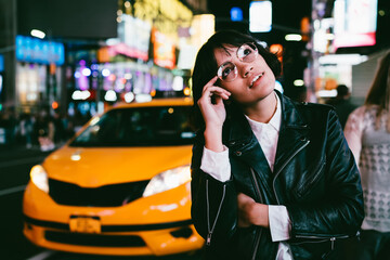 Charming brunette young woman in stylish spectacles looking up thinking at urban setting of night city,pensive hipster girl in leather jacket pondering about nightlife in metropolis standing at street