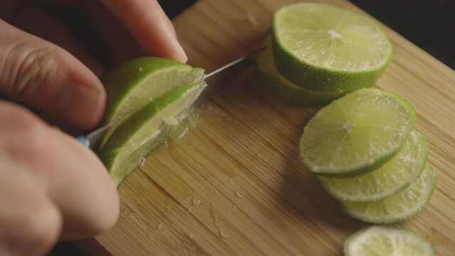 Hand Firmly Holding A Lime Fruit While Cutting It Into Thin Slices Using A Sharp Kitchen Knife - Closeup Shot 