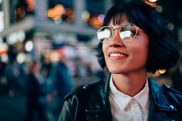Charming positive brunette young woman in cool spectacles enjoying night street standing on bokeh...