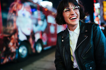 Cheerful hipster girl in design spectacles laughing feeling carefree on free time walking in downtown with night lights.positive young woman in stylish wear having fun in evening megalopolis