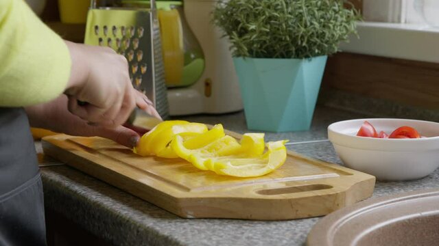 Attractive caucasian female housewife in apron slices yellow sweet paprika with a sharp kitchen knife on oak board. Cooking. Healthy eating concept. Fresh vegetables.