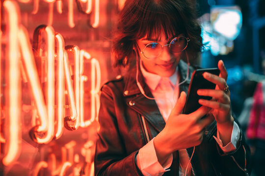Pensive hipster girl in spectacles watching video on smartphone satisfied with internet connection resting in night city, charming young woman reading message on mobile chatting in networks outdoors
