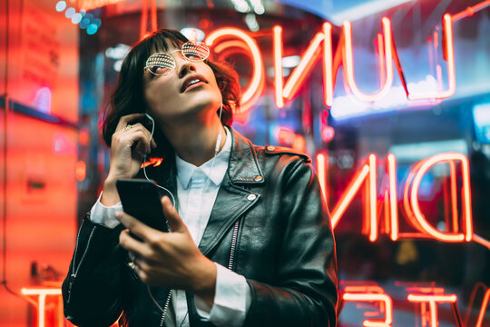 Excited female lover of music dressed in stylish leather jacket listening songs online in earphones connected to smartphone while enjoying night lights and neon illumination in New York City