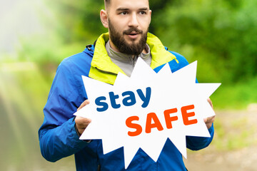 A young man in a blue jacket stands in the nature in summer and holds the message on the banner stay safe. The concept of social distance in the fight against pandemic 2020. coronavirus