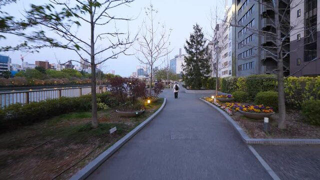 Walk at empty alley, nice small linear park at bank of Okawa river, evening time. First person view camera, aged woman slowly go ahead. Residential buildings on right