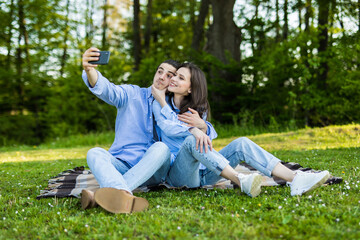 Adorable young couple man and woman sitting on green grass in park and taking selfie on smartphone