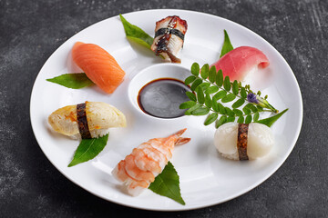 Sushi nigiri with tuna, salmon, shrimp, scallop, eel, omelet, on a white plate with soy sauce, chopsticks, leaves, on concrete background