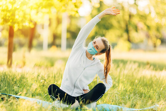 Photo of a beautiful young athlete in a medical protective mask, in a Park, doing a stretch
