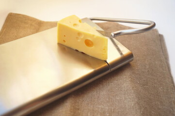 Cheese on metal cheese cutter. Metal cheese slicer with handle. On the background of a craft textile tablecloth, close-up. On white background in horizontal format and in natural light. 