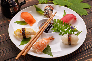 Sushi nigiri with tuna, salmon, shrimp, scallop, eel, omelet, on a white plate with soy sauce, chopsticks, leaves, on a wooden background