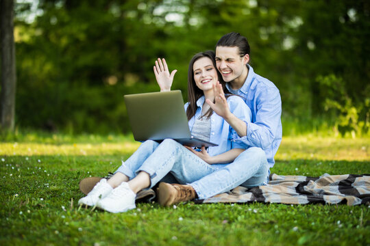 Young in loved couple with laptop make video call greeting with hands sitting on grass in park