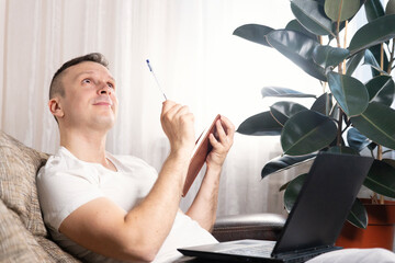 A focused European male freelancer takes notes studies working with a laptop, a young professional writes an essay in a notebook prepares for a test exam and sits at home on the couch. Remote work