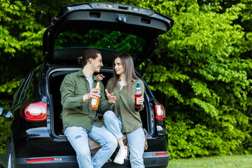 Young couple sitting together in car trunk and drinking beer in forest outdoors