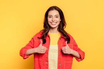 cheerful young woman showing thumbs up and looking at camera on yellow