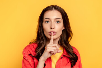 Fototapeta premium young woman showing hush sign and looking at camera on yellow