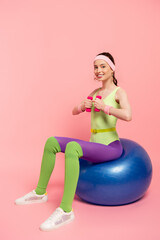 positive sportswoman exercising with dumbbells and sitting on fitness ball on pink