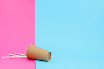 Brown paper cup inverted flipped over on pink-blue background with paper drinking straws. Disposable ecologically clean dishes.