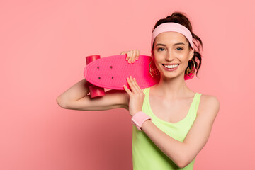 cheerful girl with headband holding penny board on pink