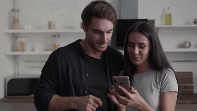The couple in their kitchen look together at the smartphone and have fun talking, they gesticulate and smile