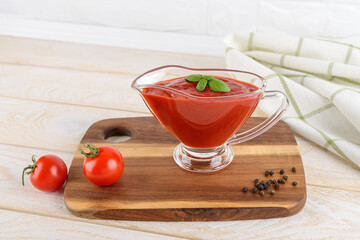 Tasty thick tomato ketchup in a glass gravy boat and ripe red cherry tomatoes on a wood cutting board over white kitchen table. Spices, seasonings and sauces.