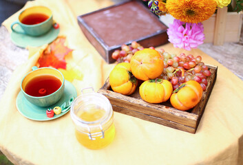 Autumn picnic. Tea party with beautiful kettle, cups at wooden table in garden. Harvest festival. Honey with stick, spoon, apple pie, persimmons, grapes, maple leaf, flowers, yellow linen tablecloth