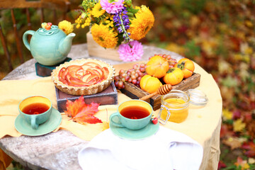 Autumn picnic. Tea party with beautiful kettle, cups at wooden table in garden. Harvest festival. Honey with stick, spoon, apple pie, persimmons, grapes, maple leaf, flowers, yellow linen tablecloth