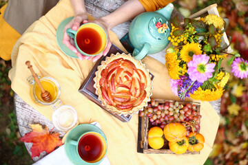 Autumn picnic. Tea party with beautiful kettle, cups at wooden table in garden. Harvest festival. Honey with stick, spoon, apple pie, persimmons, grapes, maple leaf, flowers, yellow linen tablecloth