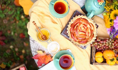 Autumn picnic. Tea party with beautiful kettle, cups at wooden table in garden. Harvest festival. Honey with stick, spoon, apple pie, persimmons, grapes, maple leaf, flowers, yellow linen tablecloth