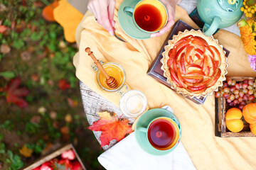 Autumn picnic. Tea party with beautiful kettle, cups at wooden table in garden. Harvest festival. Honey with stick, spoon, apple pie, persimmons, grapes, maple leaf, flowers, yellow linen tablecloth