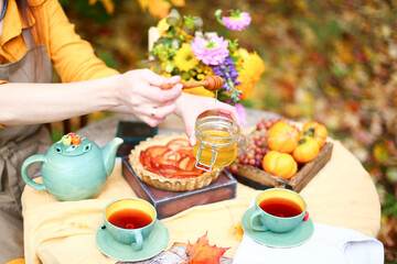 Autumn picnic. Woman in yellow dress and linen apron drinks tea from cup at wooden table in garden. Beautiful kettle, tablecloth, honey with spoon, apple pie, harvest, persimmon, grapes, maple leaf